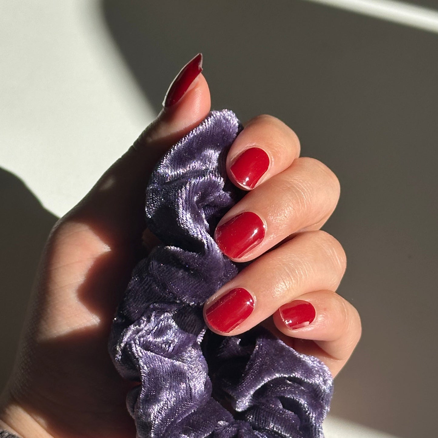Hand holding a purple scrunchie with red nail polish on a neutral background