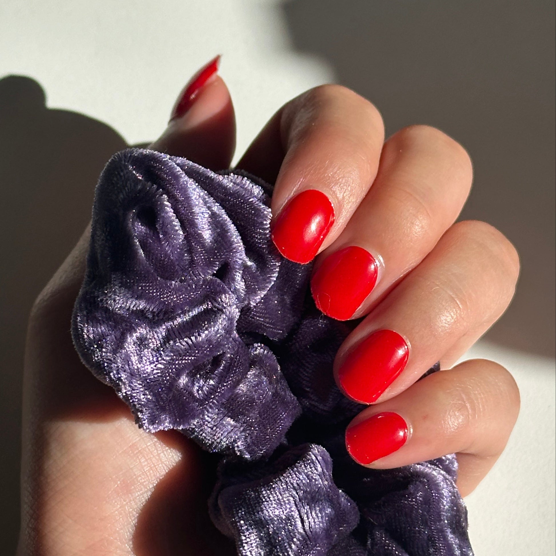 Hand holding two purple velvet scrunchies with red nail polish on a neutral background