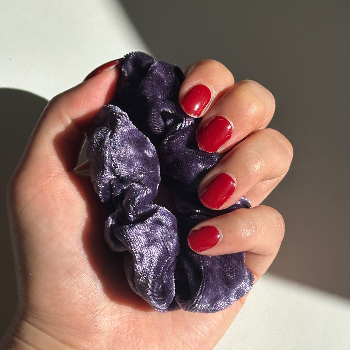 Hand holding a purple scrunchie with red nail polish on a neutral background